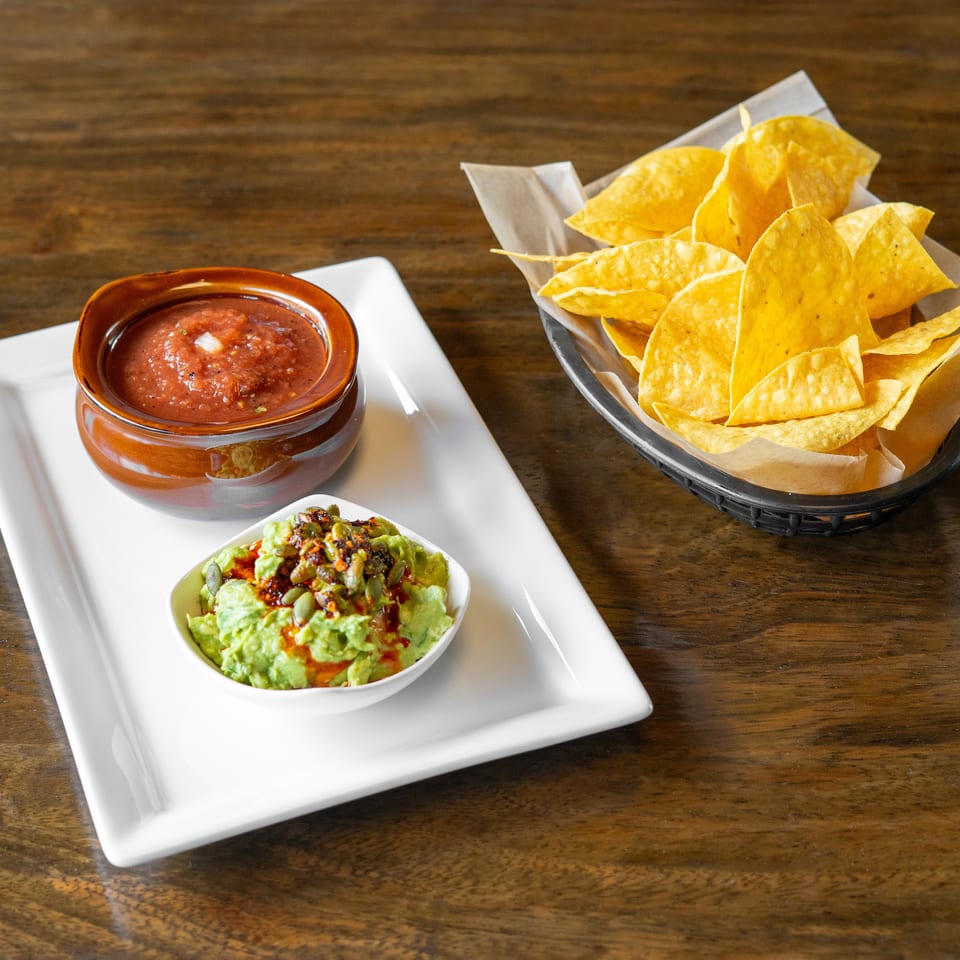 Salsa, guacamole with pumpkin seeds, and a basket of tortilla chips on a wooden table.