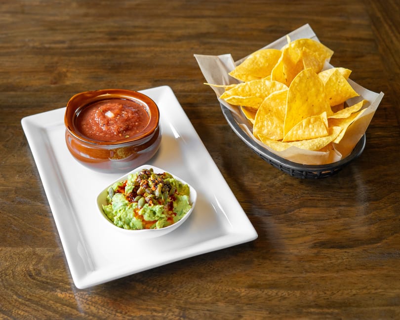 Salsa, guacamole with pumpkin seeds, and a basket of tortilla chips on a wooden table.