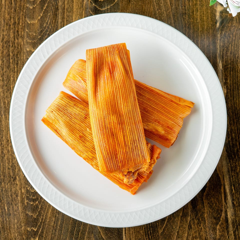 Three orange tamales, still in their corn husk wrappers, arranged on a white plate.