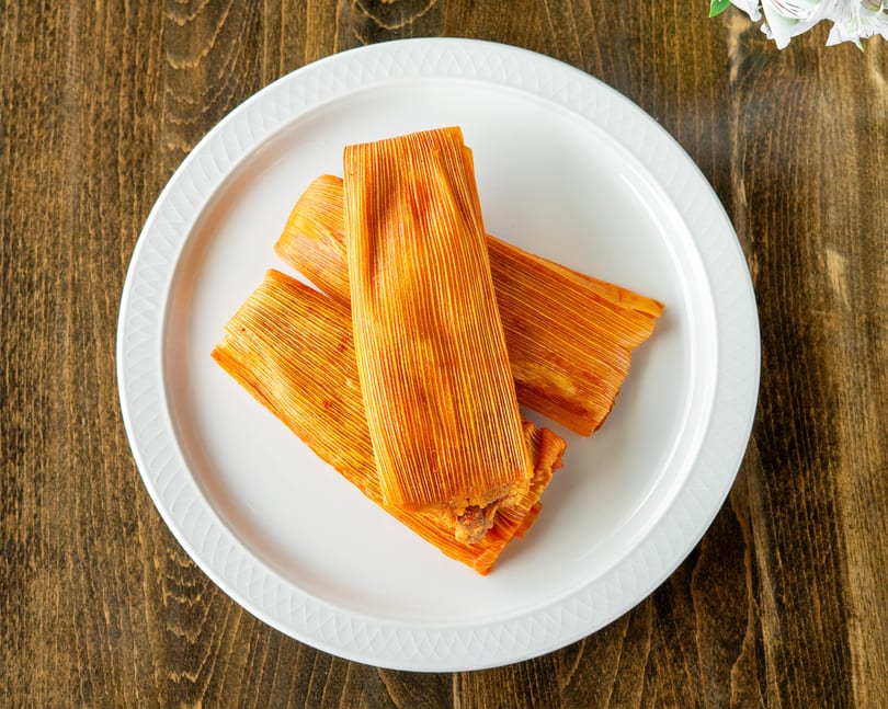 Three orange tamales, still in their corn husk wrappers, arranged on a white plate.