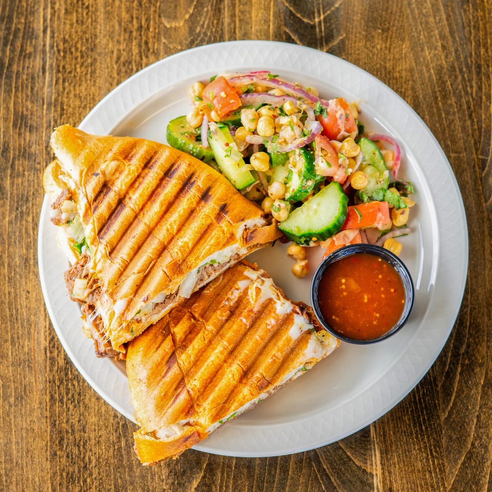 Grilled panini sandwich with melted cheese, a side salad of chickpeas, cucumbers, tomatoes, and a small cup of sauce.