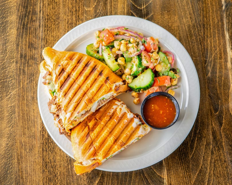 Grilled panini sandwich with melted cheese, a side salad of chickpeas, cucumbers, tomatoes, and a small cup of sauce.