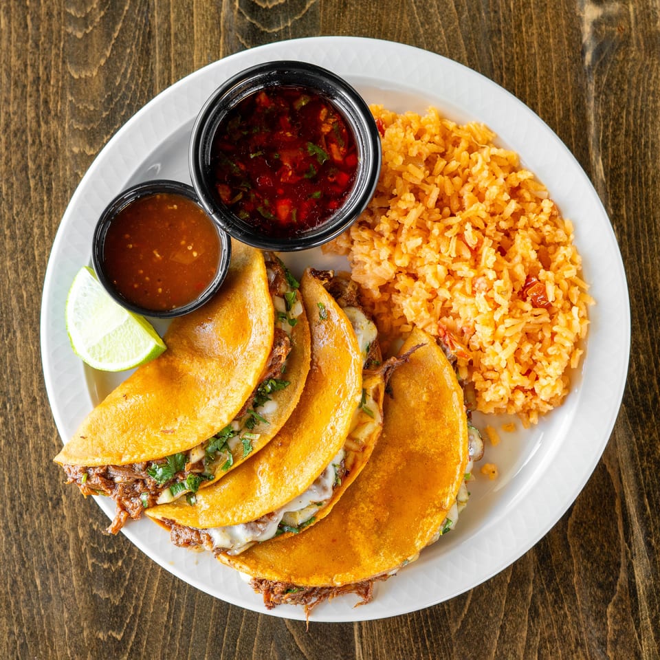 Four birria tacos with melted cheese and cilantro, served with rice, two sauces, and a lime wedge.