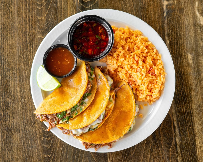 Four birria tacos with melted cheese and cilantro, served with rice, two sauces, and a lime wedge.