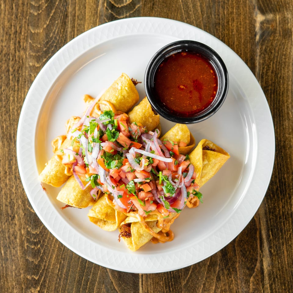 Crispy taquitos topped with pico de gallo and red onions, served with a side of red sauce.