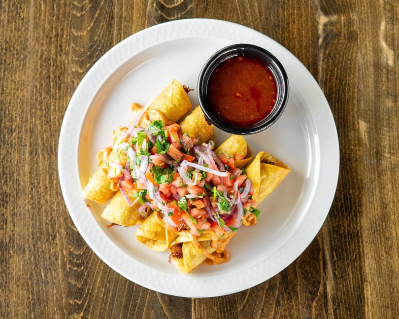 Crispy taquitos topped with pico de gallo and red onions, served with a side of red sauce.