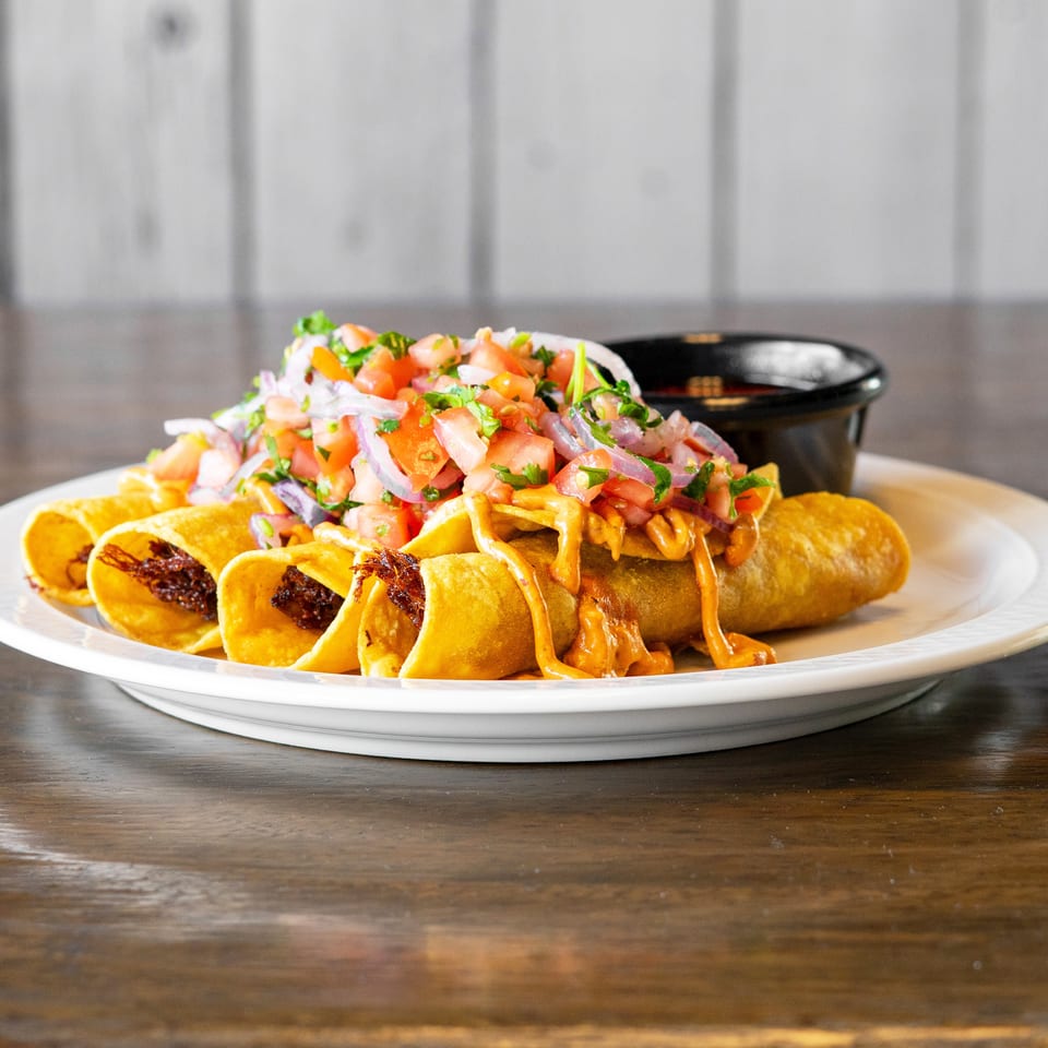 Crispy taquitos topped with fresh pico de gallo and a creamy orange sauce, served with a side of salsa.