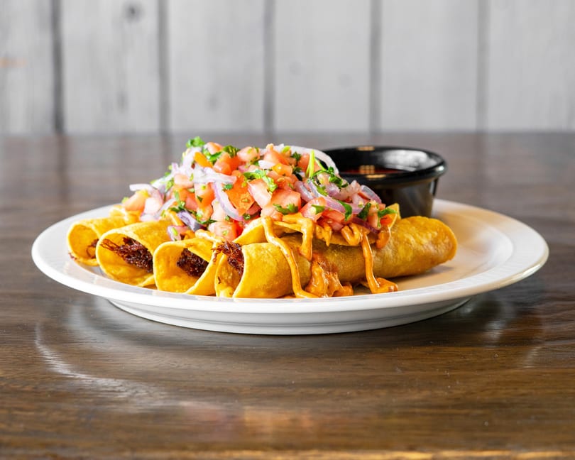 Crispy taquitos topped with fresh pico de gallo and a creamy orange sauce, served with a side of salsa.