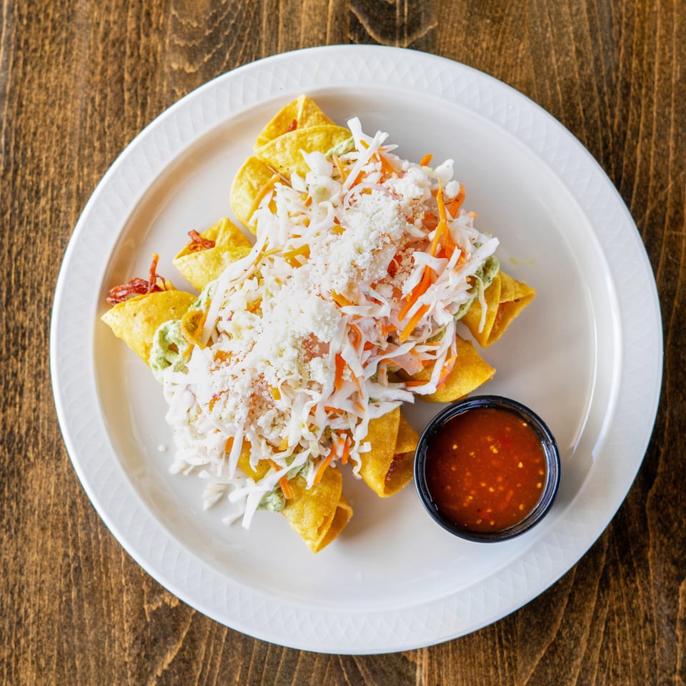Golden taquitos topped with shredded cabbage, carrots, cheese, and a side of red salsa.