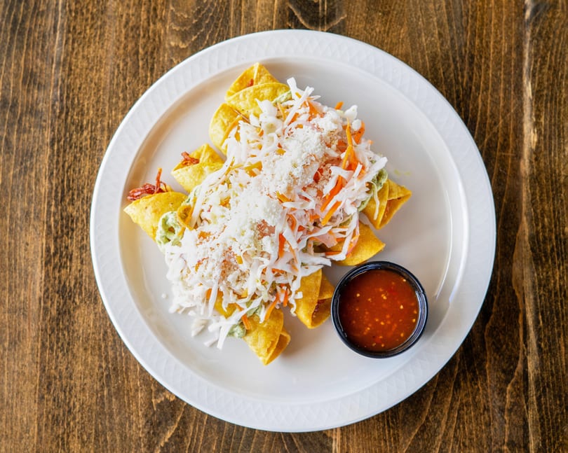Golden taquitos topped with shredded cabbage, carrots, cheese, and a side of red salsa.
