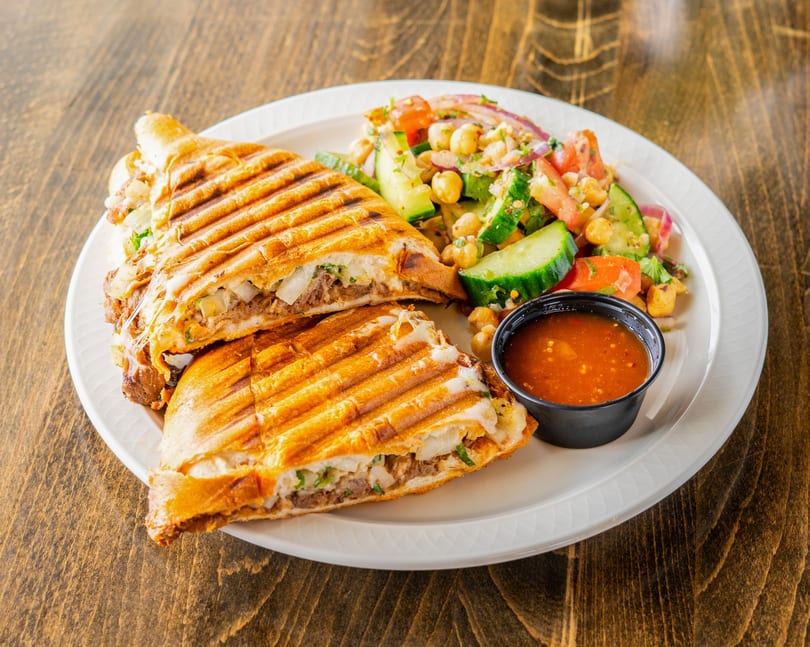 Grilled panini sandwich with meat and cheese, served with a side salad and dipping sauce.